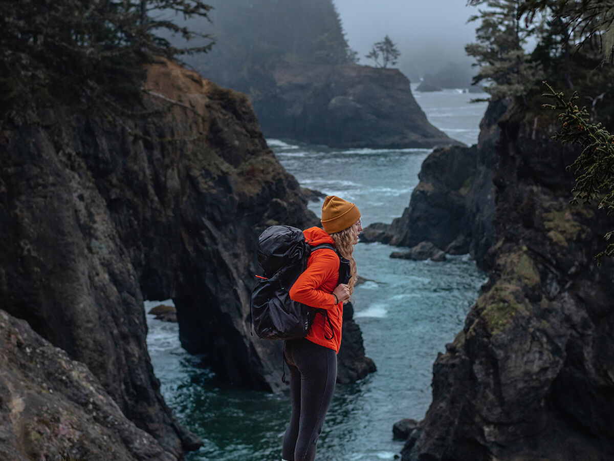 A person stands on a cliff edge, wearing a backpack and winter clothing, overlooking a misty coastal landscape with jagged rocks and trees.