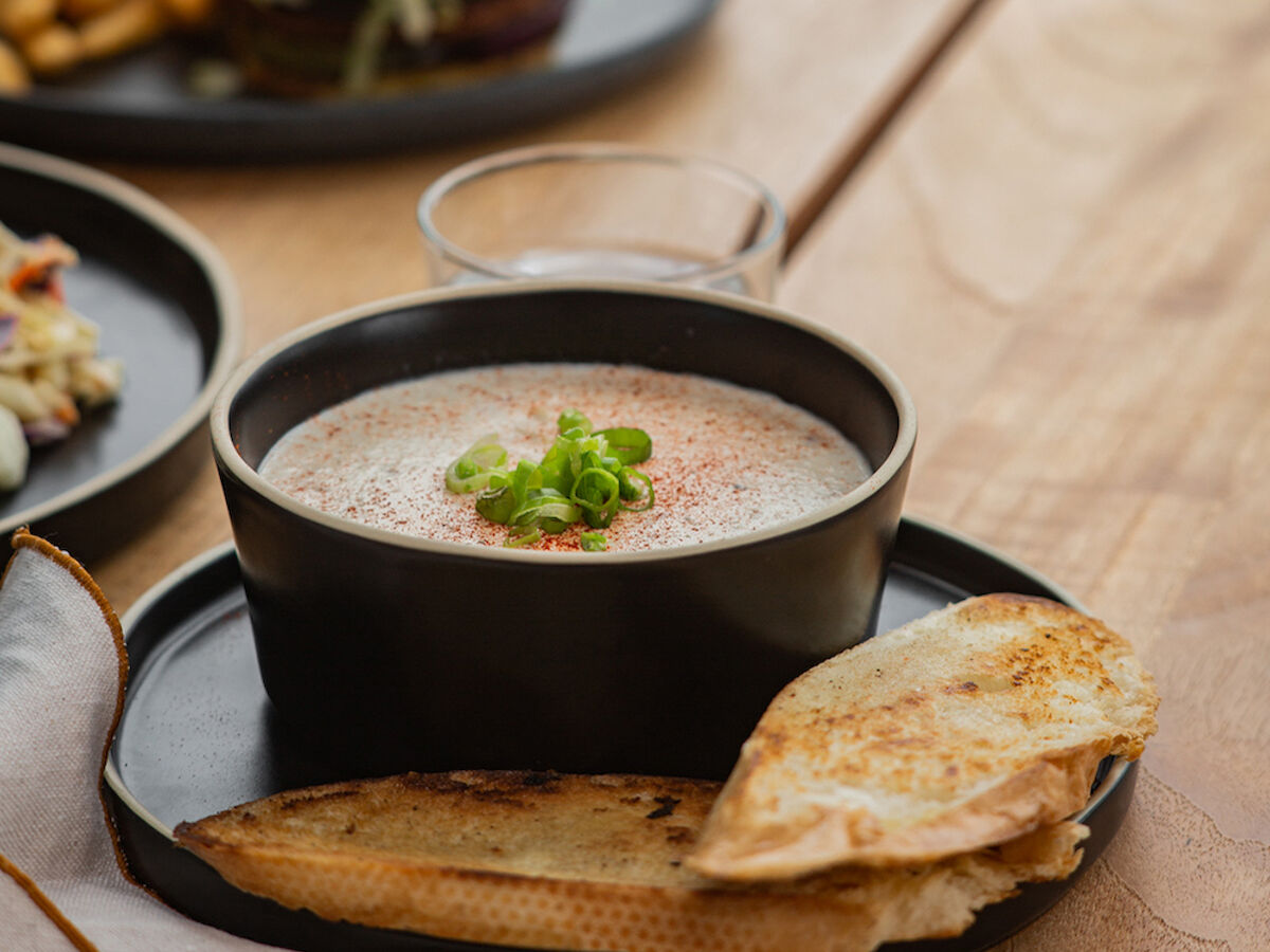 A bowl of creamy soup garnished with herbs and served with toasted bread on a wooden table; a burger and fries in the background.