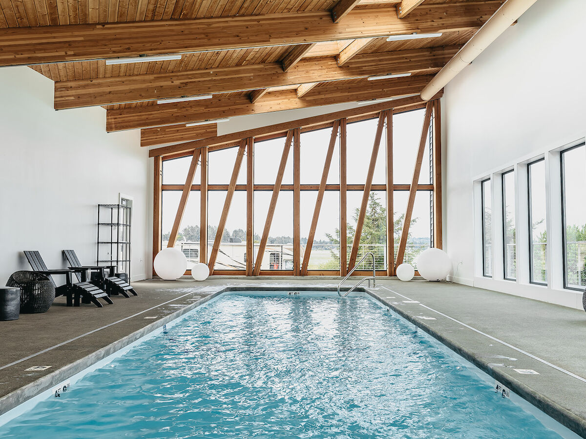 Indoor pool with modern design, angled wooden ceiling, large windows, and lounge chairs beside the blue water, creating a relaxing space.