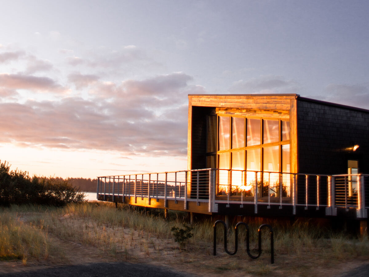 A modern building with large windows, reflecting the sunset, stands against a partly cloudy sky, surrounded by a natural landscape.