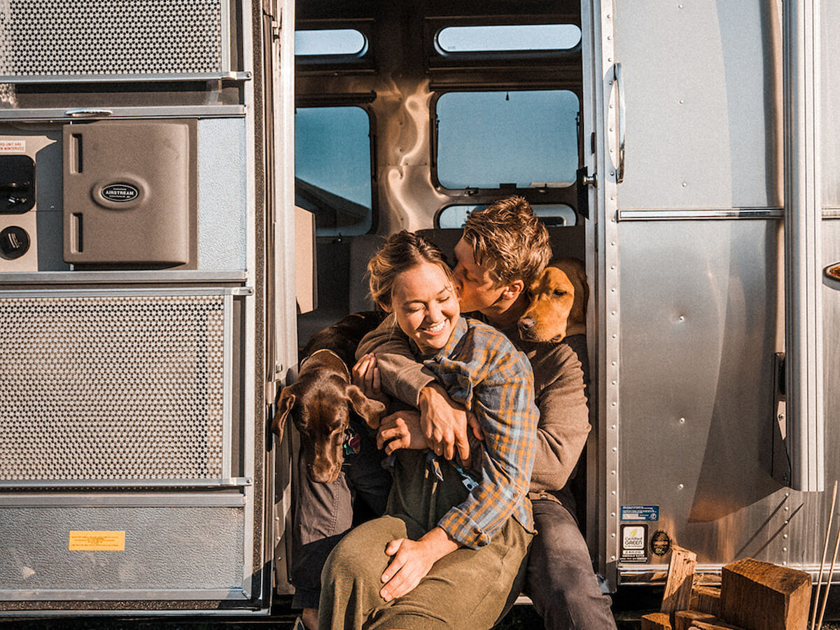 A couple sits on the steps of a camper van, cuddling a dog, with a relaxed and joyful expression on their faces.