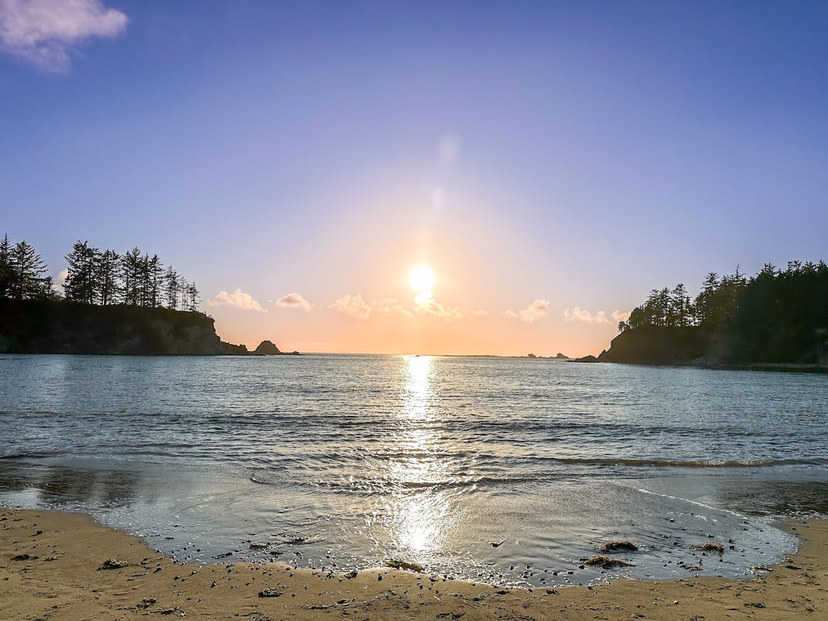 A serene beach scene with a clear sky, calm waves, and the sun setting on the horizon. Trees silhouette the edges of the coastline.