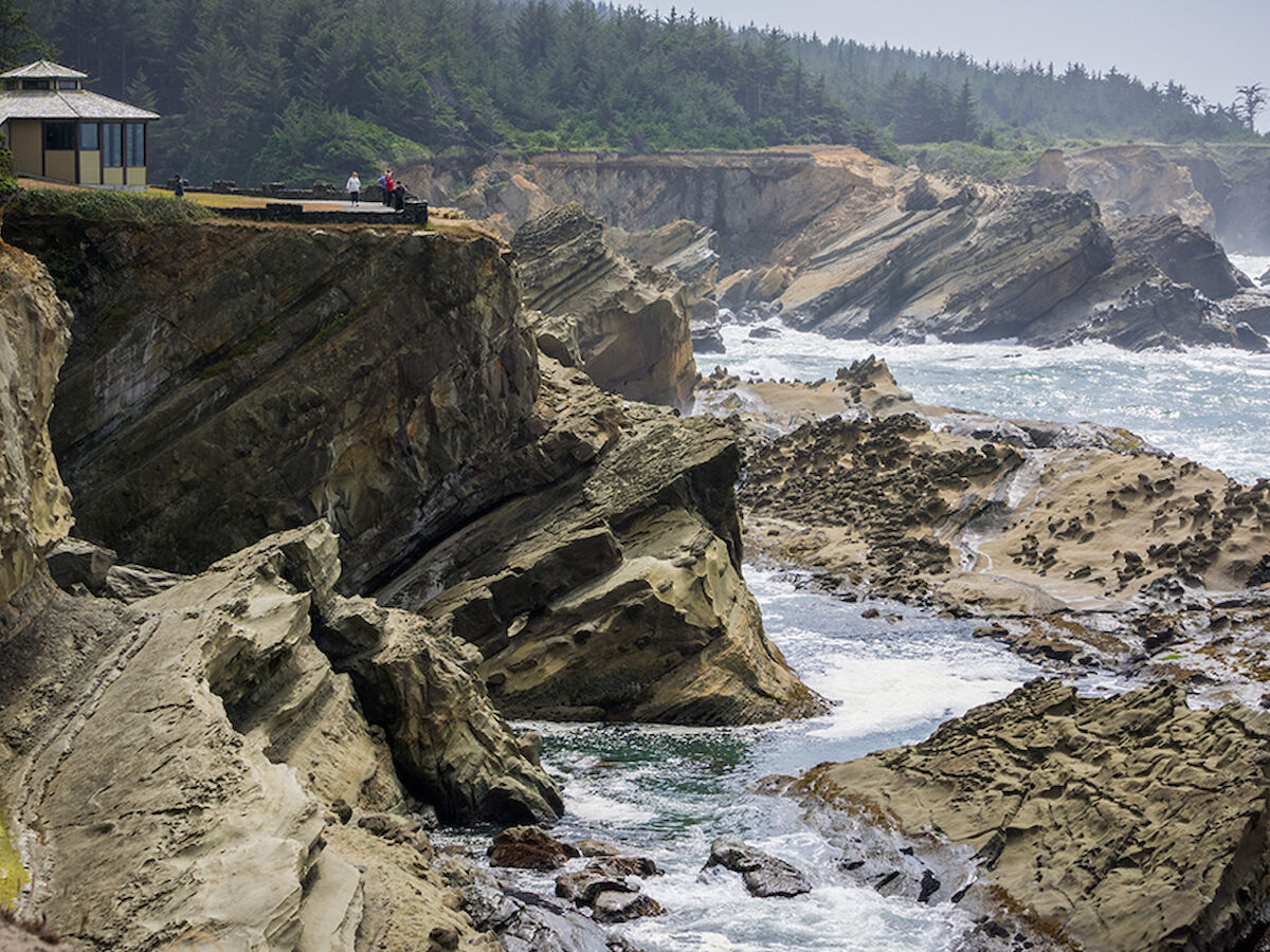 A scenic view of rugged cliffs meeting the ocean, with a small structure perched on the edge and dense forest in the background.