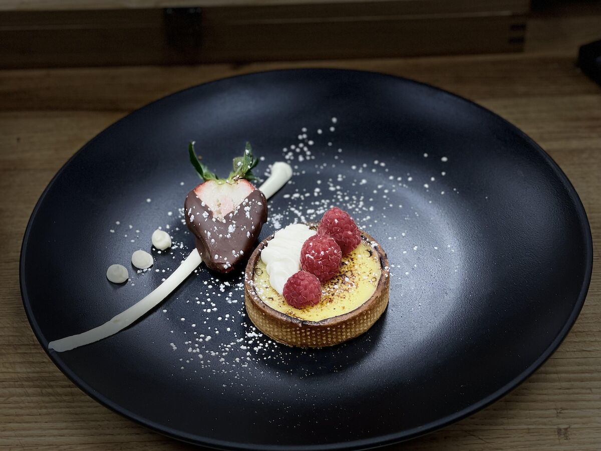 A dessert plate featuring a tart topped with cream and raspberries, accompanied by a chocolate-covered strawberry, and artistic sauce decor.