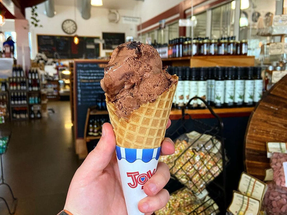 A person holds a chocolate ice cream cone in a waffle cone inside a shop, surrounded by various products and shelves.