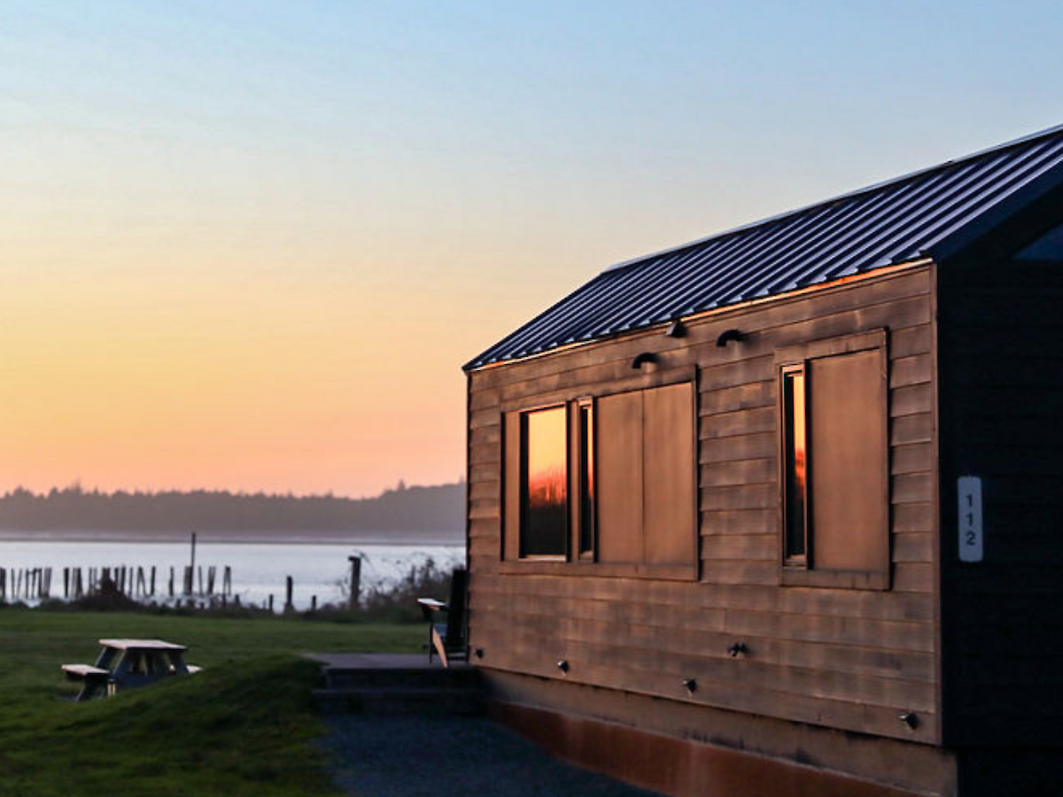 A small wooden cabin near a lake or river at sunset. A picnic table is nearby, creating a peaceful and serene outdoor setting.