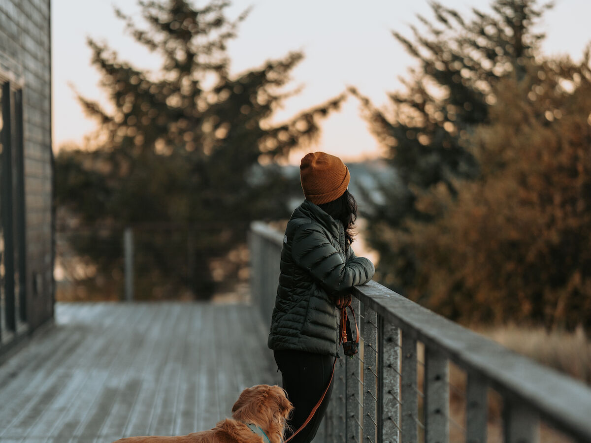 A person in a beanie stands on a wooden deck with a dog, facing trees and the Oregon Coast.