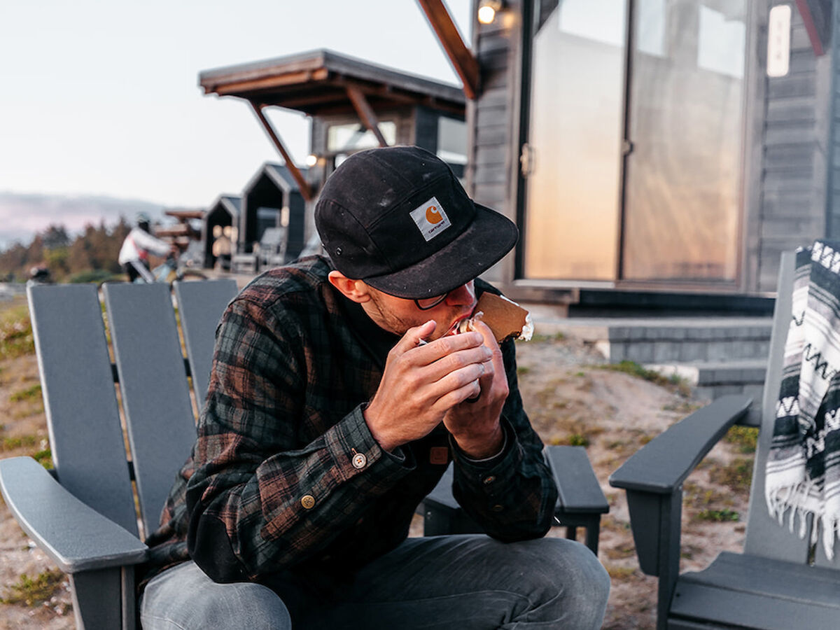 A person sits outside, drinking from a mug, near modern cabins and outdoor seating, capturing a cozy outdoor moment.