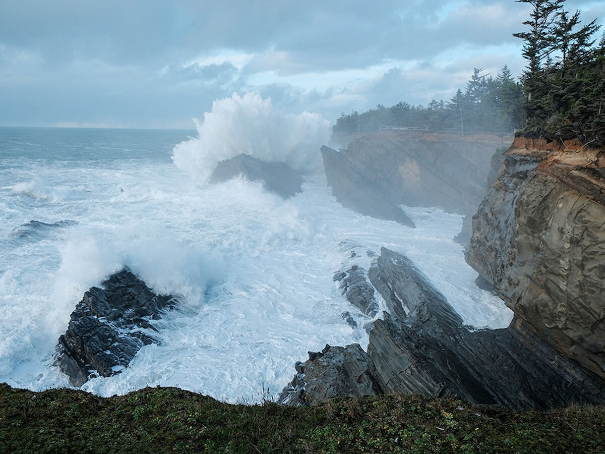 Waves crash against rocky cliffs under a cloudy sky, surrounded by trees and a rugged coastline.