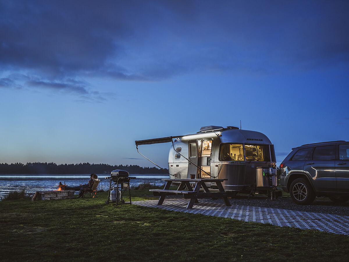 A camper trailer with an awning is parked by a lake, near a picnic table and a barbecue, at dusk.