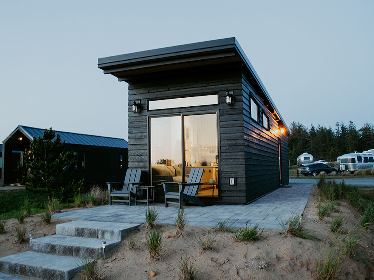 A modern tiny house with a patio and chairs in a grassy area, surrounded by other tiny homes and RVs at dusk.