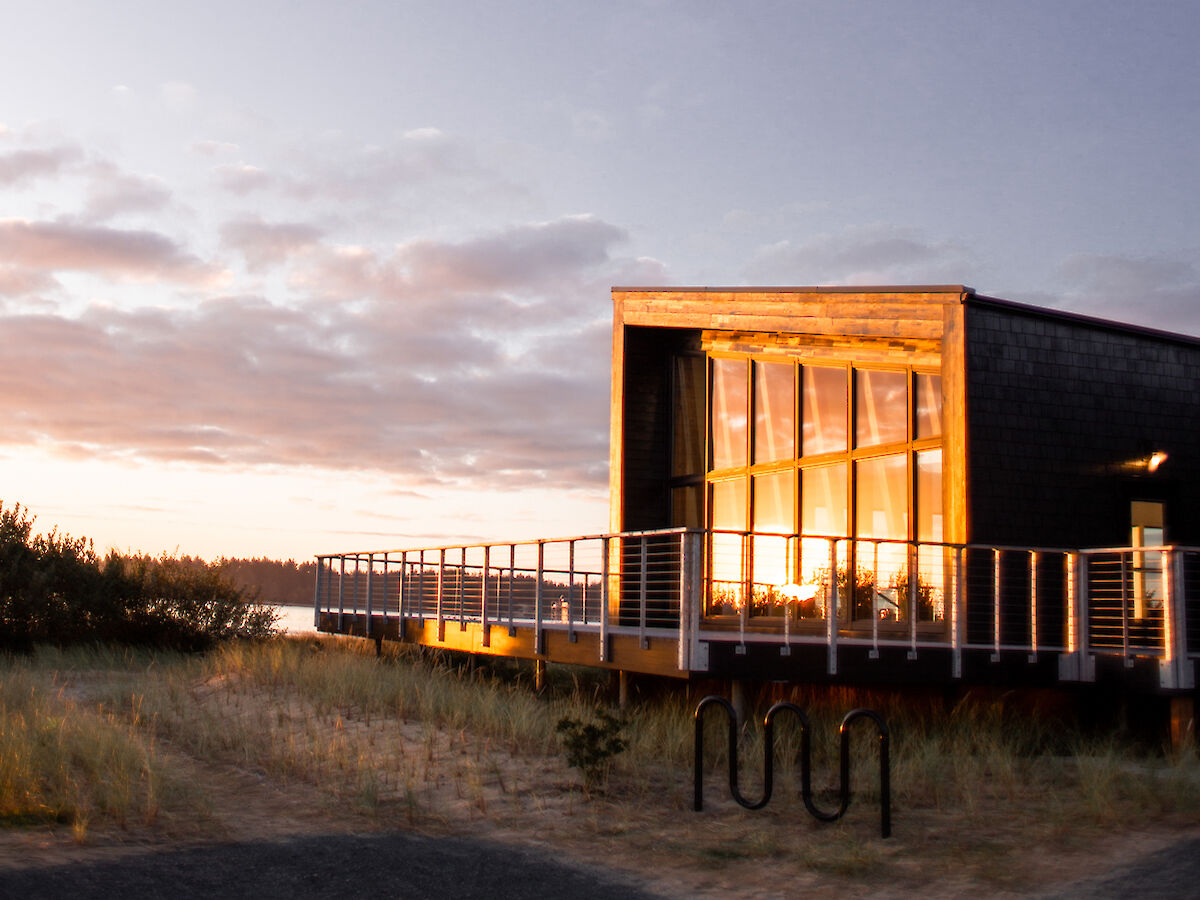 A modern building with large windows is lit by the setting sun, surrounded by a natural landscape under a clear sky.