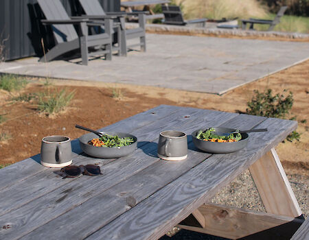 A modern cabin with outdoor seating, a wooden picnic table set with cups and plates, and a scenic view of water and landscape in the background.