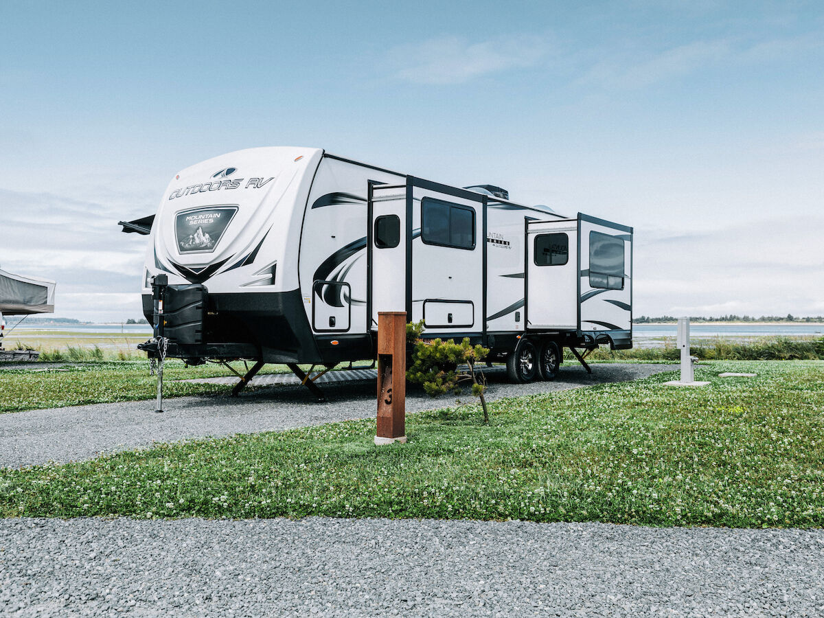 The image shows a white travel trailer parked on a grassy area with a clear sky backdrop.