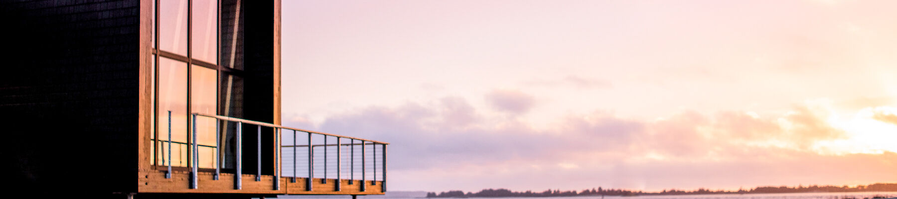 A modern cabin with large windows sits by a serene beach at sunset, reflecting warm colors across a tranquil landscape.