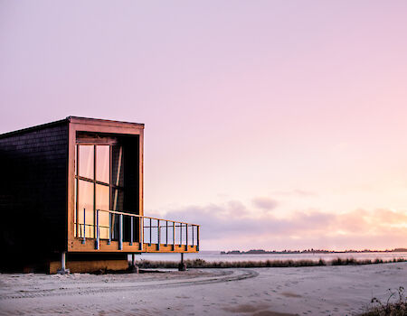 A modern cabin with large windows sits by a serene beach at sunset, reflecting warm colors across a tranquil landscape.