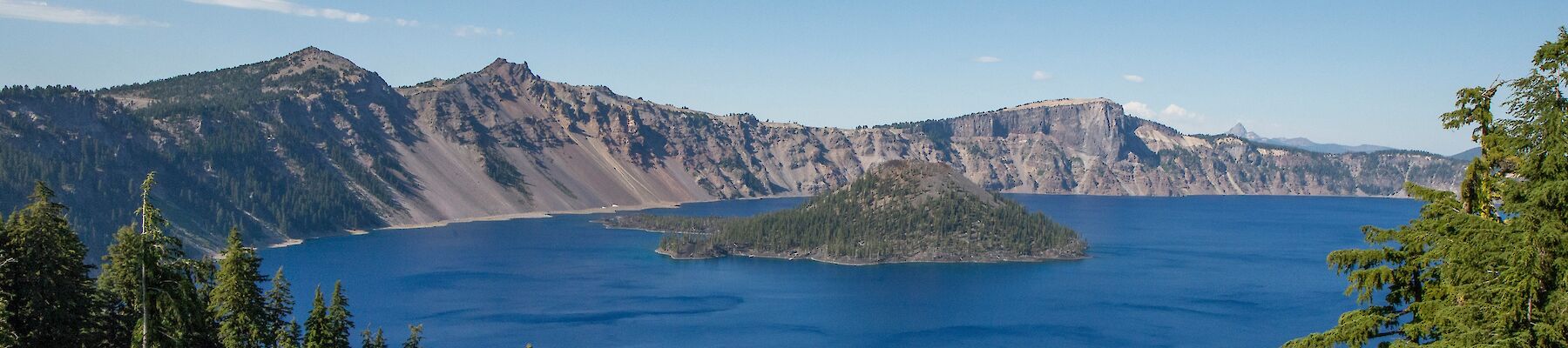 A scenic view of a deep blue lake surrounded by rugged mountains and dense pine forests, with a small island in the lake centered.