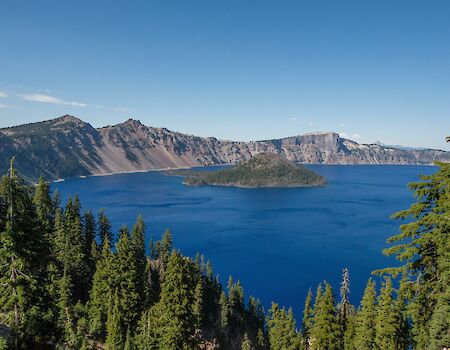 A scenic view of a deep blue lake surrounded by rugged mountains and dense pine forests, with a small island in the lake centered.