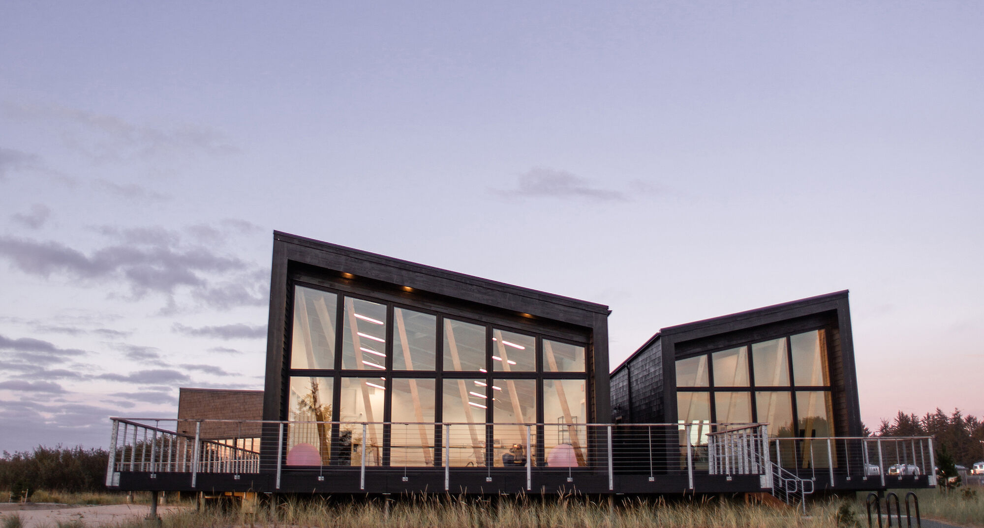 A modern building with large glass windows and a slanted roof set against a clear sky at dusk, surrounded by grass and some trees.