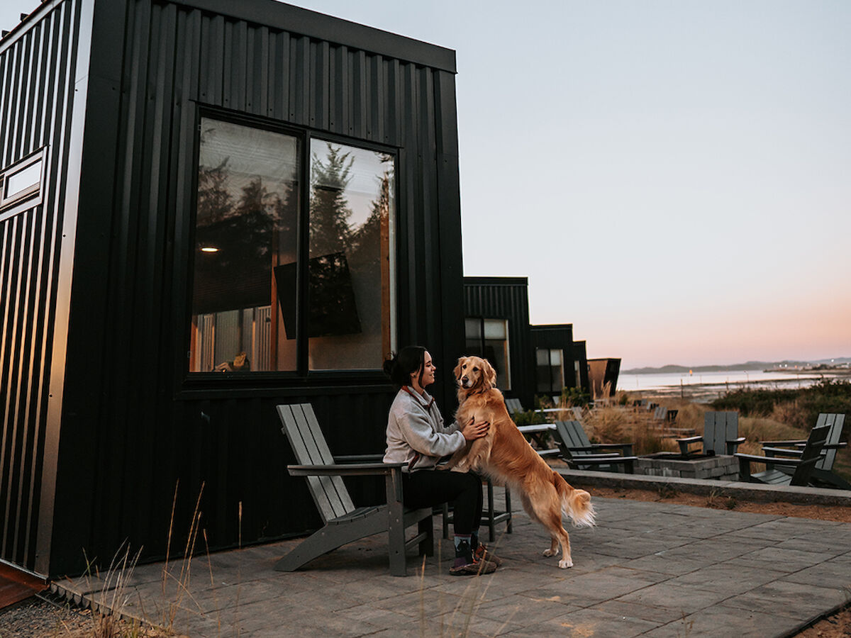 A person sits outside a small black modern cabin, petting a standing dog, with more cabins and a scenic landscape in the background.