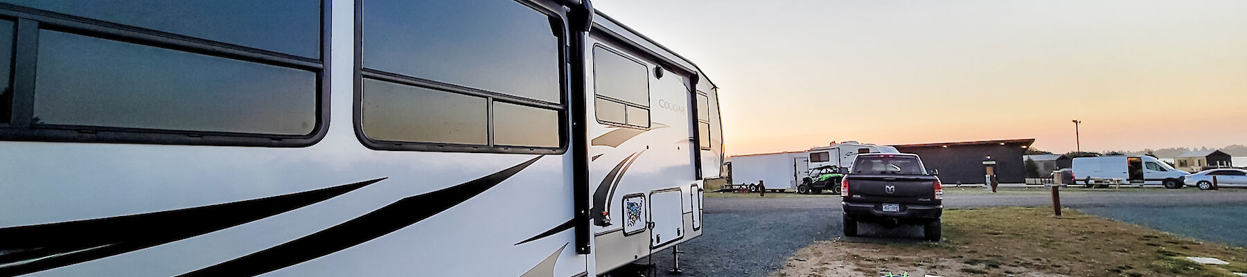 A camper and truck are parked on gravel near a picnic table with a bucket, set against a clear sunset sky.