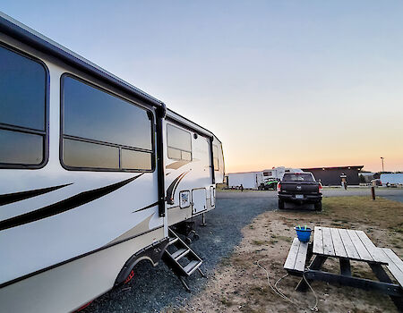 A camper and truck are parked on gravel near a picnic table with a bucket, set against a clear sunset sky.