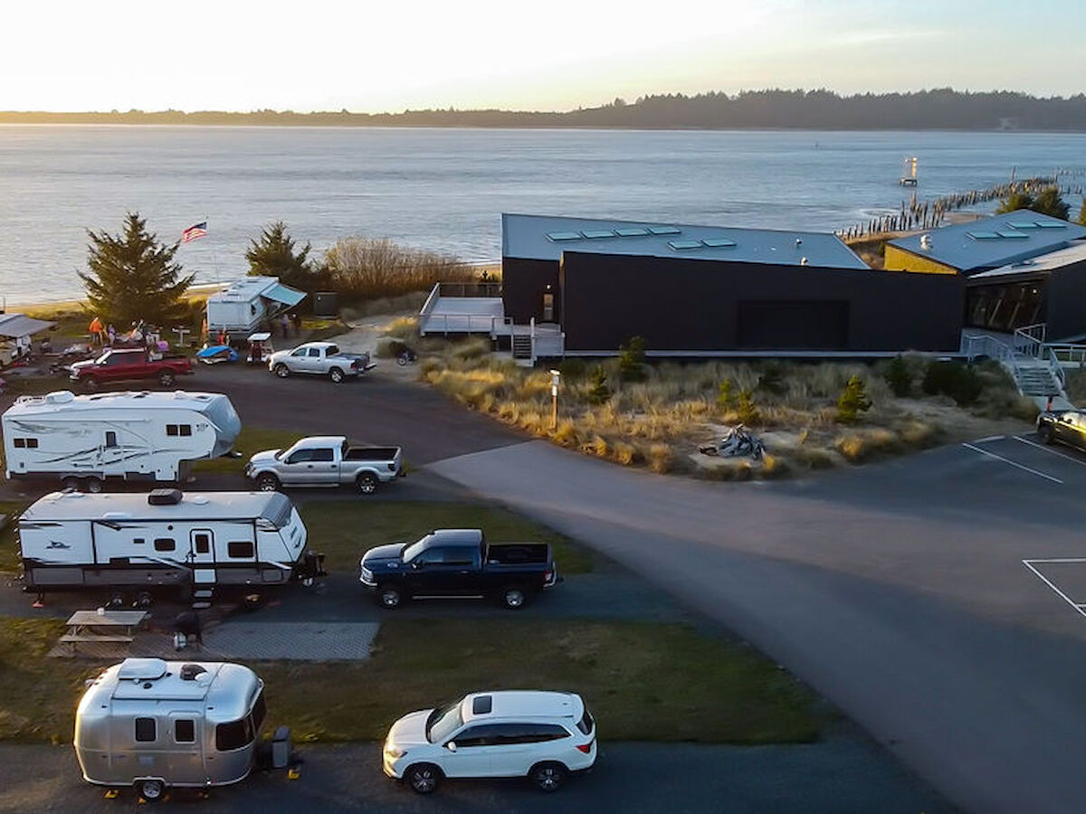 The image shows a waterfront area with several RVs, cars, and a modern building, adjacent to a body of water with trees in the background.