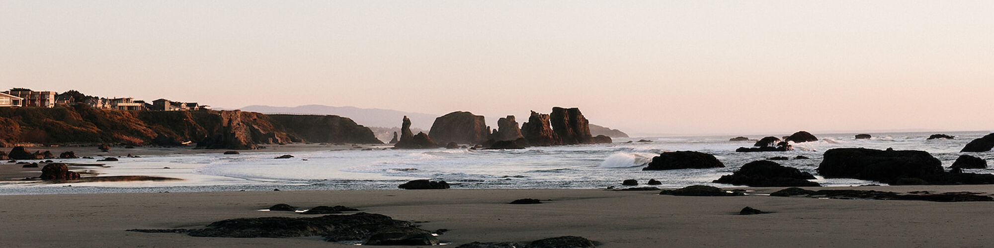 A serene beach scene with rock formations in the water near the shoreline and gentle waves; distant cliffs line the horizon of the coastline.