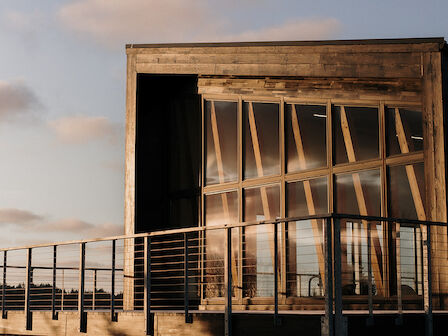 A modern, wooden beach house with large windows and a deck, set against a backdrop of sand dunes and cloudy sky.