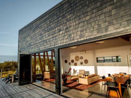 A modern house with large open windows shows a stylish interior with wooden furniture and wall decor, set against a blue sky.