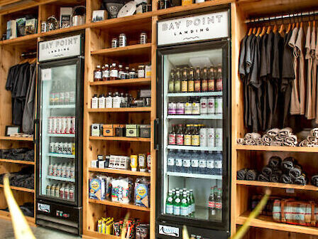 The image shows a shop interior with refrigerated displays, shelves of products, and racks of clothing. There's a plant in the foreground.