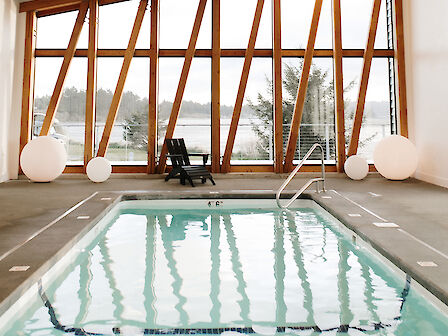 An indoor pool with modern architecture, slanted windows, wooden ceiling beams, and round lamps. There are lounge chairs by the poolside.