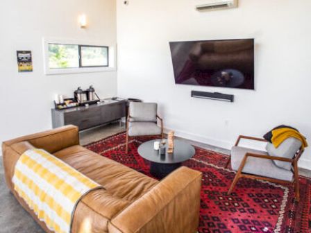 A cozy living room with a leather sofa, TV, chairs, and a patterned rug on the floor. A window provides natural light, enhancing the decor.