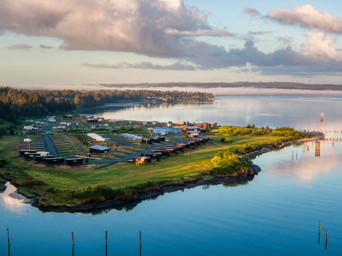 Aerial view of a coastal RV park on a small peninsula with lush greenery, surrounded by calm blue waters and a cloudy sky.