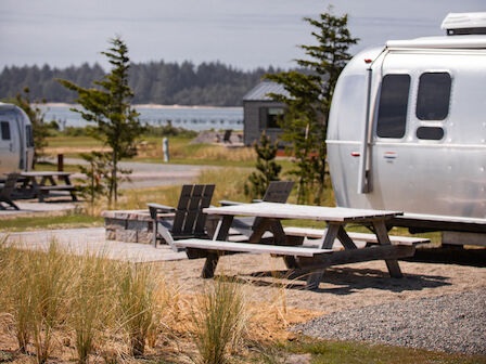 The image shows a scenic campground with two silver trailers, picnic tables, grass, trees, and a body of water under a cloudy sky.