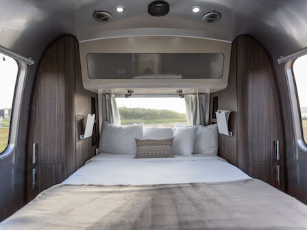 A cozy RV interior with a neatly made bed, surrounded by wooden panels and windows, offering a view of the outdoors.