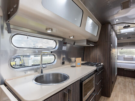 A modern RV interior with a kitchen, featuring a sink, stove, and cabinets, leading to a cozy sleeping area in the back.