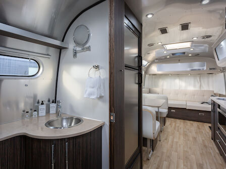 The image shows a modern camper interior with a sleek design, featuring a kitchenette, sink, seating area, and wood-style flooring.