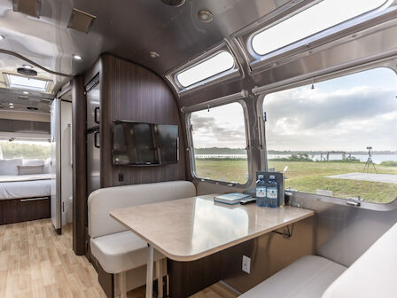 The image shows the interior of a modern RV with a dining area, sofa, and large windows offering a scenic outdoor view.