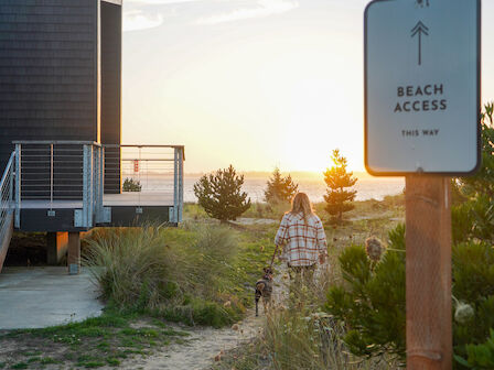 The image shows a beach access path with a sign and a person walking, set against a sunset backdrop and surrounded by natural landscape.