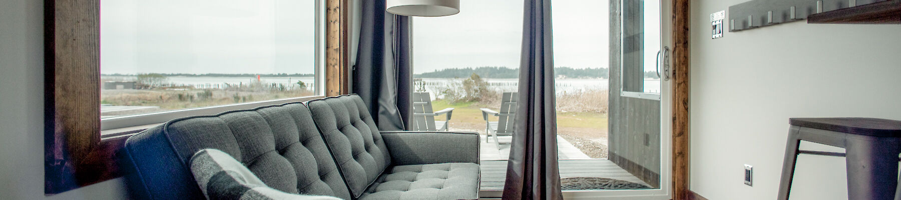 A cozy cabin room with large windows, a gray sofa, TV, and lamp. Wood accents and a view of the outdoors create a warm atmosphere.