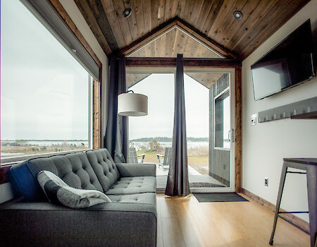 A cozy cabin room with large windows, a gray sofa, TV, and lamp. Wood accents and a view of the outdoors create a warm atmosphere.