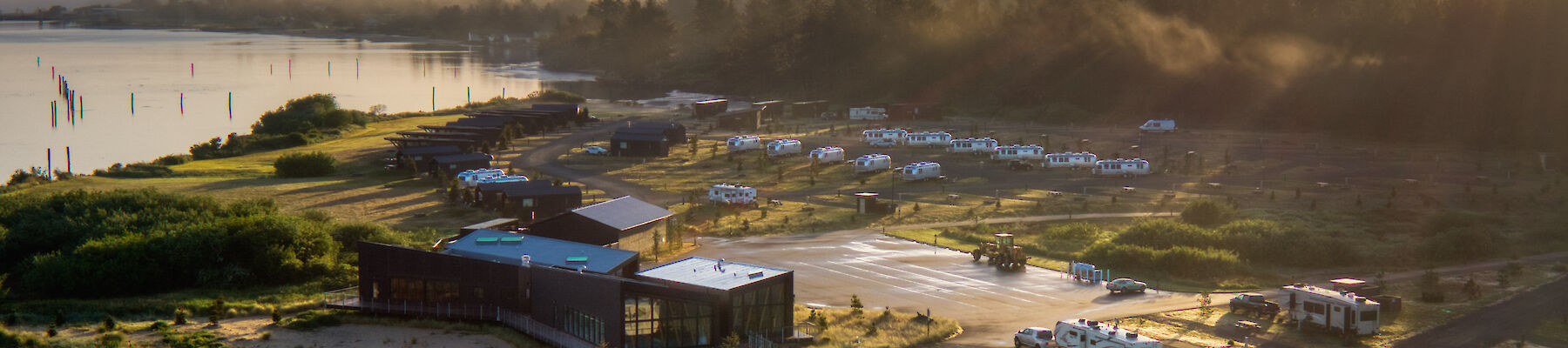 Aerial view of a campsite with RVs near a river and forest, under a sunrise or sunset sky.