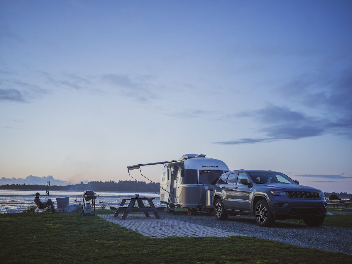 A car is parked next to a RV trailer near a picnic table. Two people sit by the Oregon Coast, enjoying the scenic view during sunset.