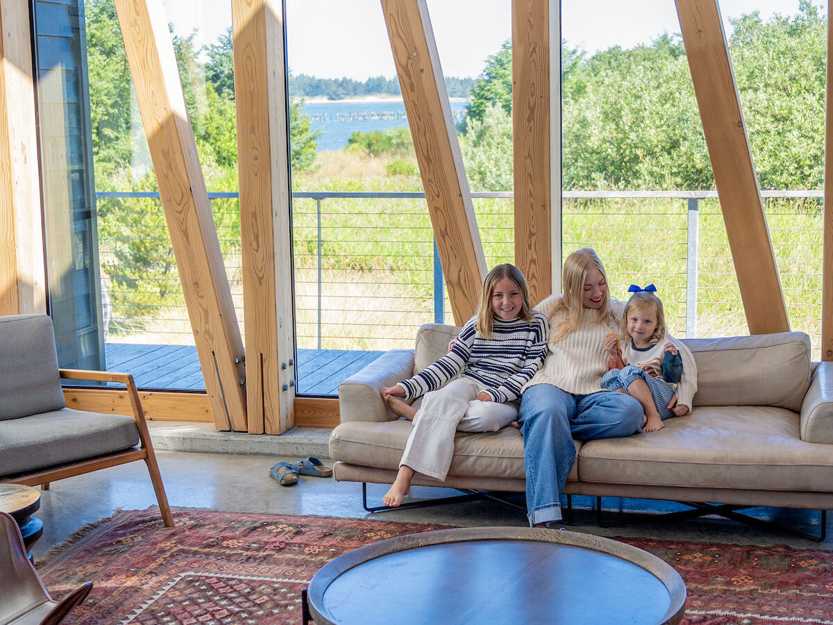 A family of three sits on a sofa in a wooden-framed room with large windows overlooking a scenic outdoor view.